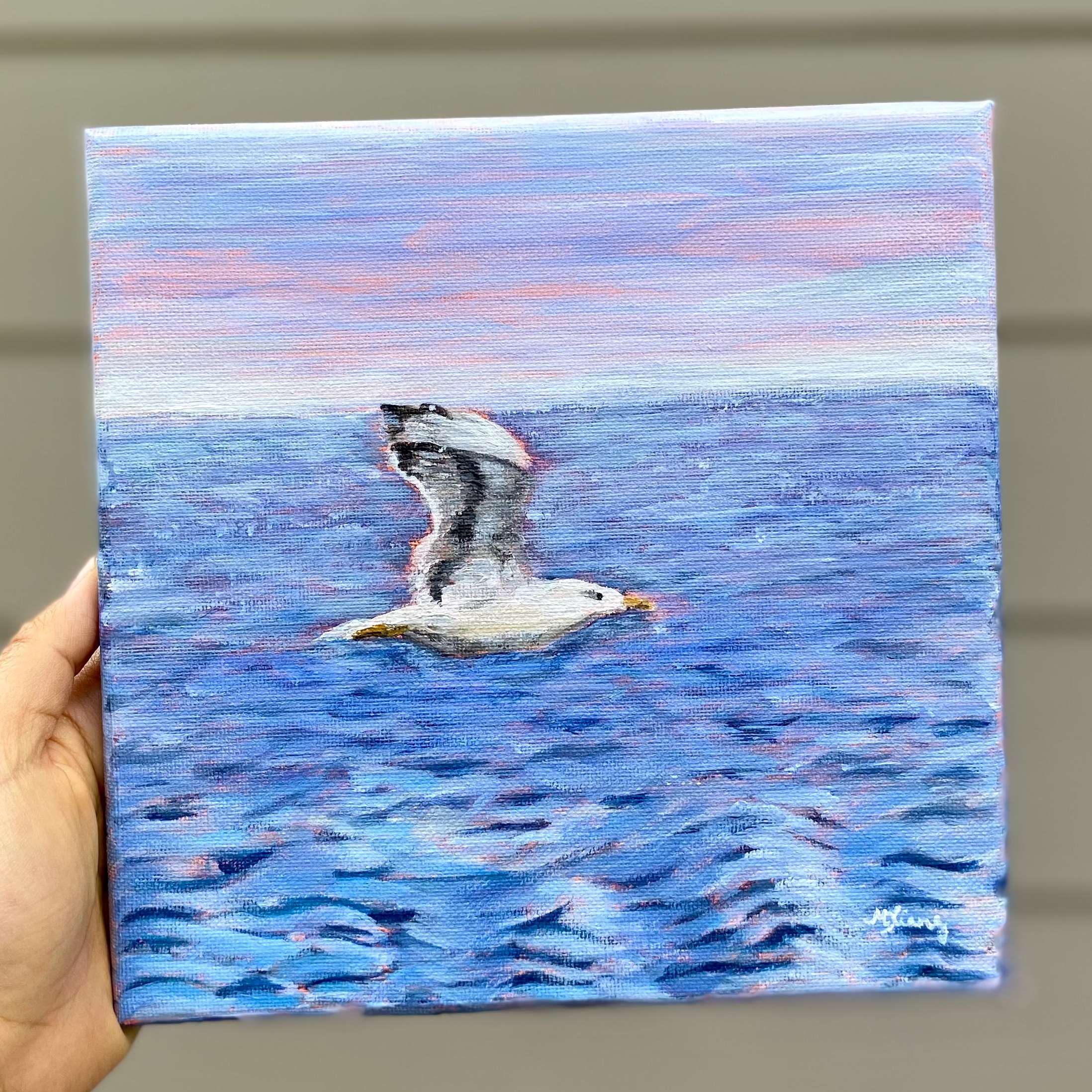 A hand holds a square canvas painting of a seagull flying over blue water under a pink and blue sky, with a beige paneled wall in the background.