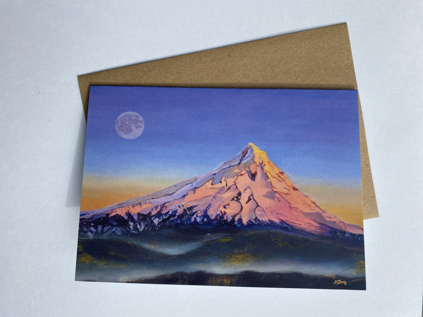 Greeting card with a mountain Mt Hood painting in sunset alpenglow glow with a full moon in the violet and orange sky, and brown envelope on a light gray background