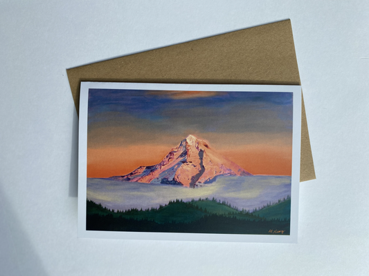 Greeting card featuring painting of a detailed mountain Mt Hood with sunset alpenglow glow with dark green tree line in the foreground and a blue and orange sky. A brown envelope on a light gray background