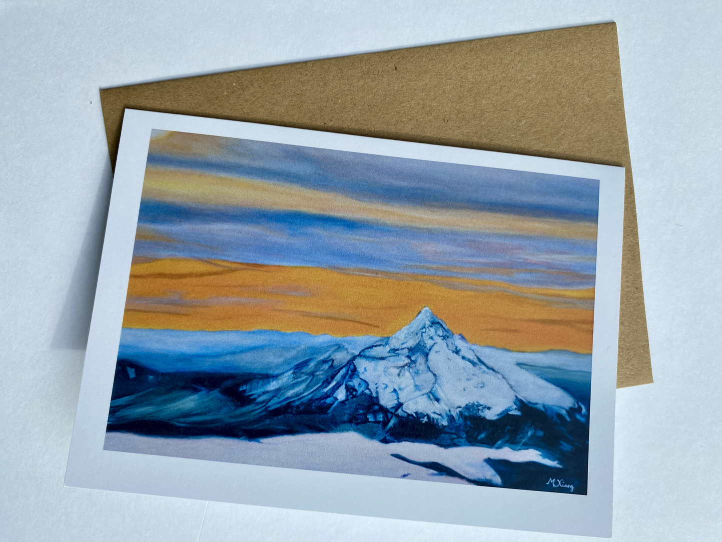Greeting card featuring painting of a detailed aerial mountain Mt Hood with sunrise glow with misty clouds and grey, orange and yellow sky. A brown envelope on a light gray background
